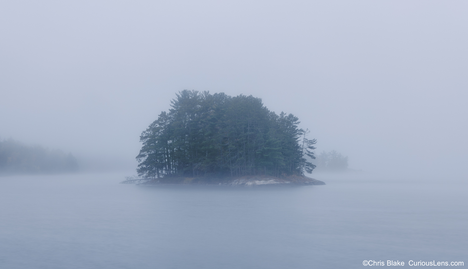 A small pine-covered island on Kabetogama Lake emerges from thick morning fog at sunrise in Voyageurs National Park, Minnesota.
