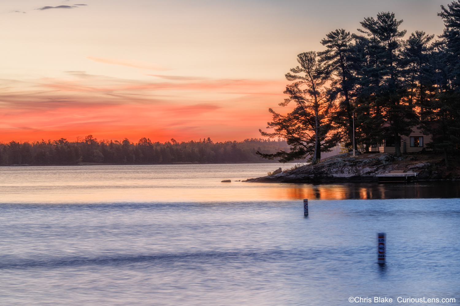 A lakeside cabin surrounded by tall pines glows in the first light of sunrise under a vivid red sky on Kabetogama Lake in Voyageurs National Park, Minnesota.
