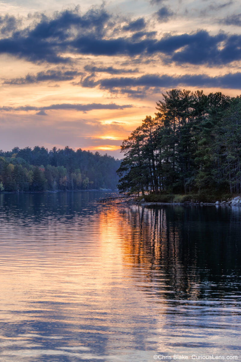 Warm evening light reflects across the calm surface of Kabetogama Lake in Voyageurs National Park, highlighting pine trees along the shoreline as the sun breaks through scattered clouds.
