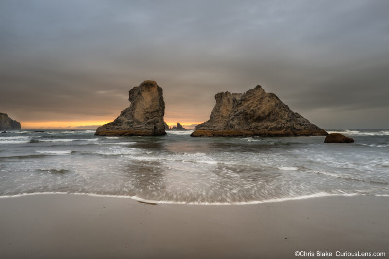 Storm clouds over large sea stacks at Bandon Beach on the Oregon Coast, with sunset color on the horizon and waves moving across the sand.
