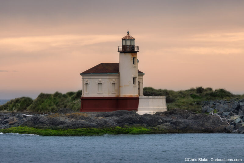Coquille River Lighthouse at sunset near Bandon, Oregon, photographed across the river with soft evening light and coastal grasses behind it.
