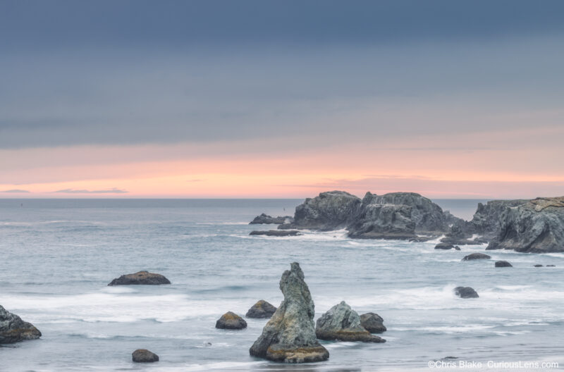 Sea stacks and ocean waves at Bandon Beach on the Oregon Coast, with soft sunset color along the horizon beneath heavy clouds.