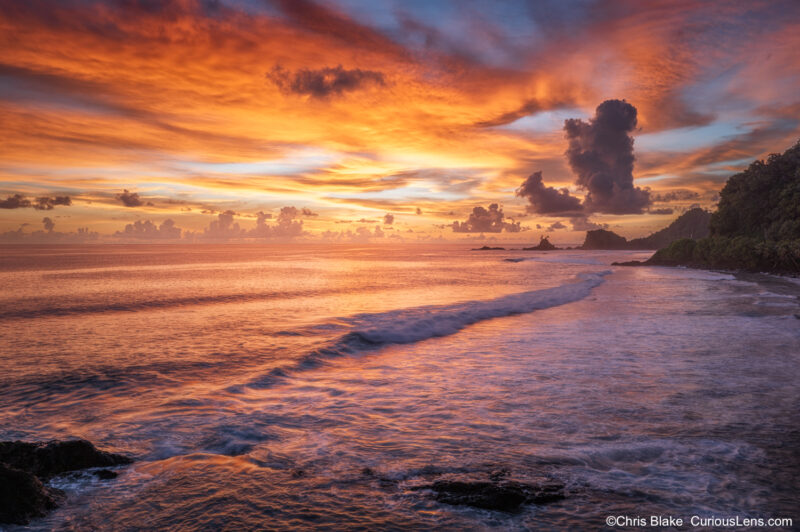 Fiery sunset over the coast of American Samoa, with glowing clouds, ocean waves, rocky shoreline, and a small sea stack with a palm tree.