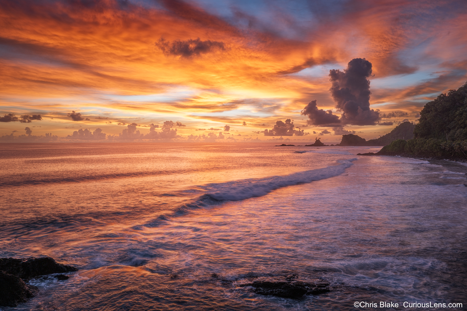 Fiery sunset over the coast of American Samoa, with glowing clouds, ocean waves, rocky shoreline, and a small sea stack with a palm tree.