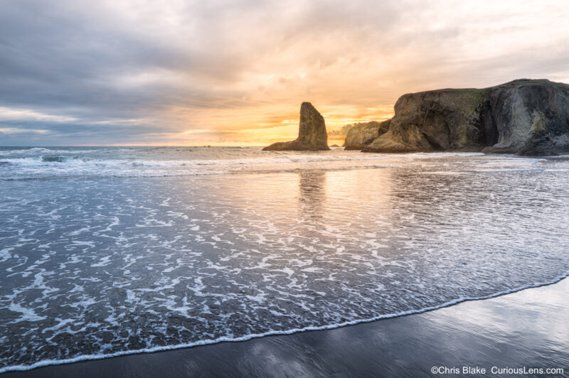 Golden sunset light over the sea stacks at Bandon Beach on the Oregon Coast, with ocean waves and reflections across the wet sand.