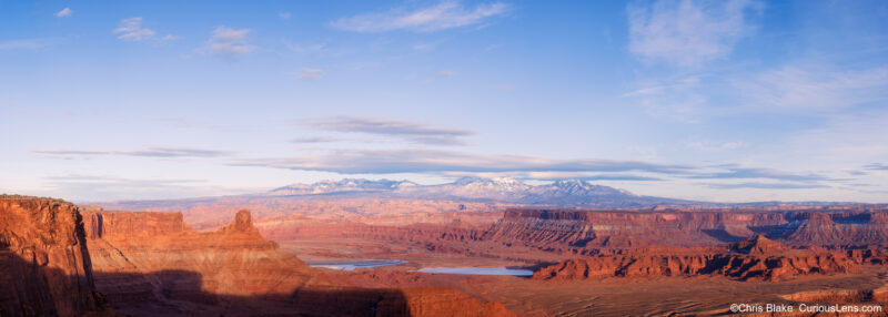 Large panoramic photograph of the La Sal Mountains from Dead Horse Point State Park in Utah, with red rock canyons, desert shadows, and late afternoon light.