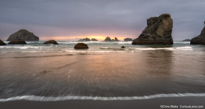 Last light at sunset breaking through storm clouds over the sea stacks at Bandon Beach, with waves rushing across the wet sand.