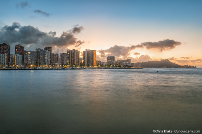 Sunrise over the Waikiki skyline and harbor in Honolulu, Hawaii, with Diamond Head in the distance and soft morning light reflecting on the ocean.
