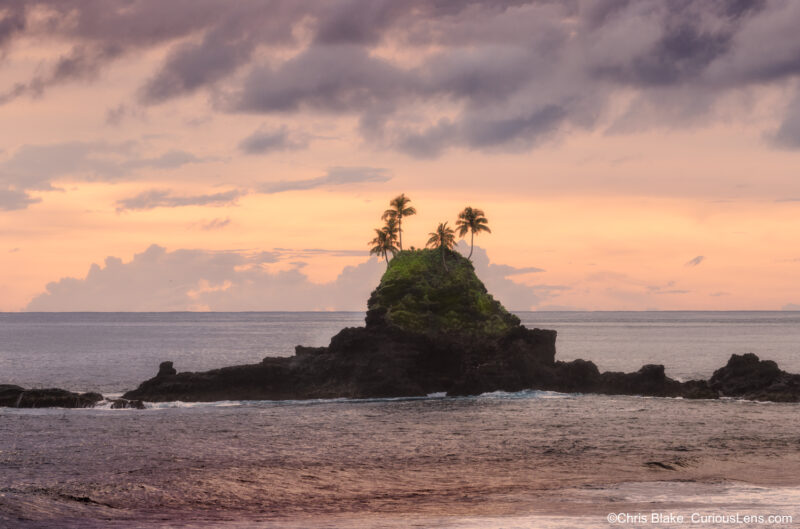 Pastel sunset over the coast of American Samoa, with a small palm-topped sea stack rising from the ocean beneath soft pink and purple evening clouds.