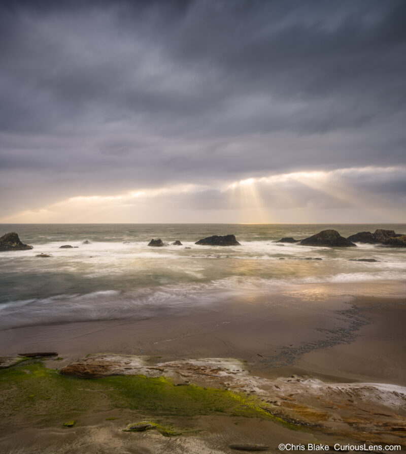 Sunlight breaking through storm clouds over Seal Rock on the Oregon Coast, with ocean waves, beach, and rain light near sunset.