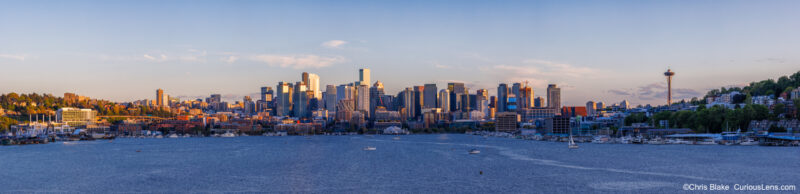 Panoramic view of the Seattle skyline across Elliott Bay from Gas Works Park, with the Space Needle, downtown buildings, and warm evening light.