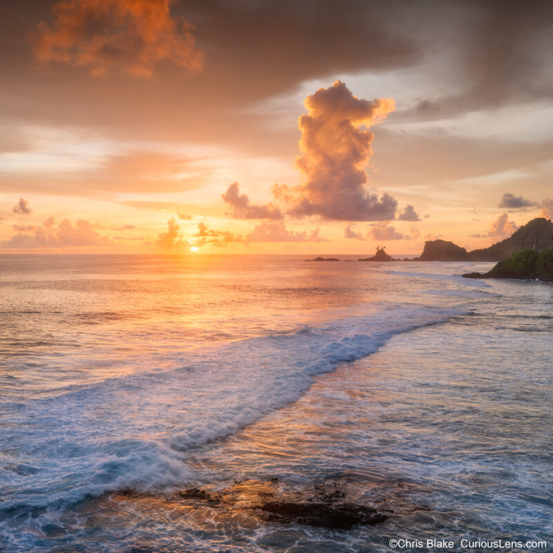 Sunset over the South Pacific coast of American Samoa, with waves rolling toward shore, glowing orange clouds, and the sun just above the ocean horizon.