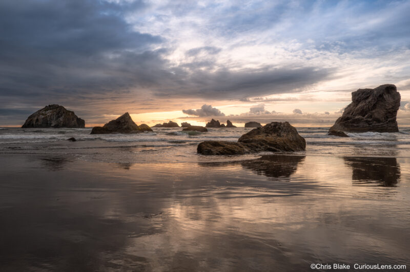 Storm clouds clearing over Bandon Beach at sunset, with sea stacks and golden reflections in the wet sand as the tide moves out.