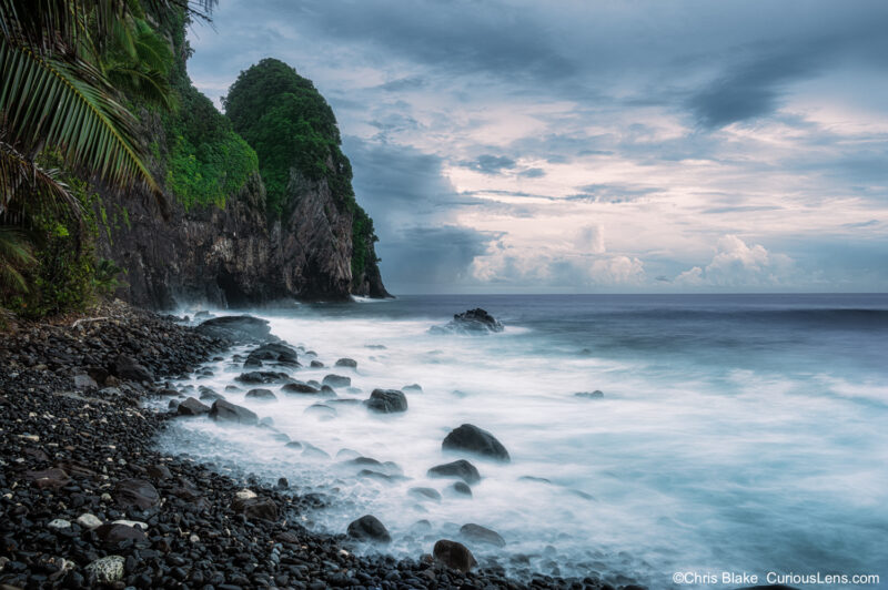 Stormy sunrise at Pola Island Beach in American Samoa, with a rocky shoreline, lush green cliffs, and long-exposure waves moving beneath dark morning clouds.