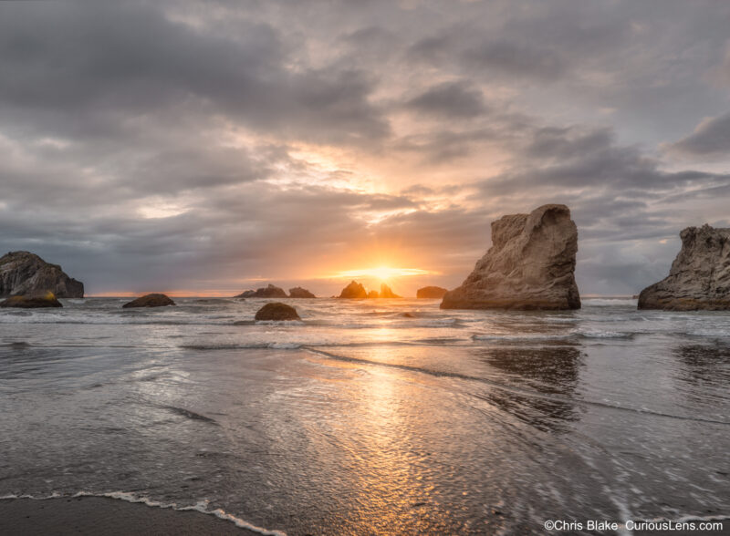 Sunset breaking through clouds between the sea stacks at Bandon Beach, with reflected golden light across the wet sand and shallow water.
