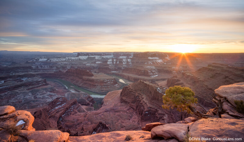 Sunset at Dead Horse Point State Park in Utah, with a sunstar over the canyon, the Colorado River below, snow in the valley, and warm light on red sandstone and desert juniper.