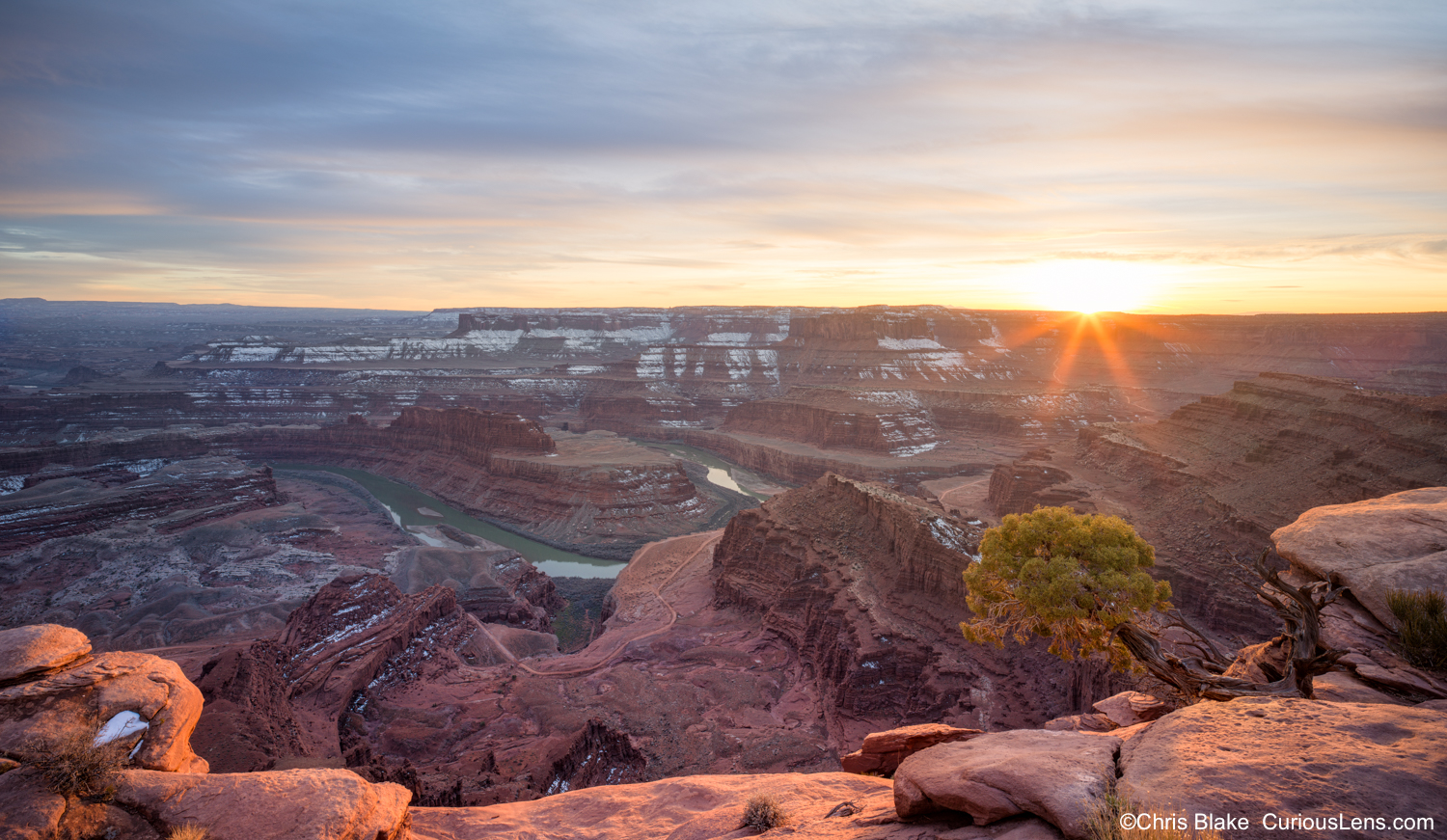 Sunset at Dead Horse Point State Park in Utah, with a sunstar over the canyon, the Colorado River below, snow in the valley, and warm light on red sandstone and desert juniper.