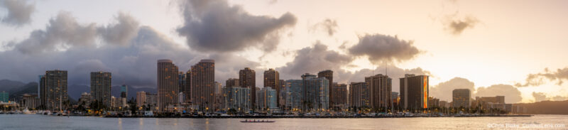 Panoramic sunrise photograph of the Waikiki skyline in Honolulu, Hawaii, seen from Magic Island with harbor water, city buildings, clouds, and rowers in the foreground.