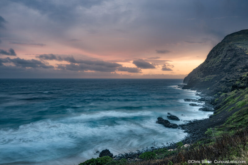 Stormy sunrise along the windward coast of Oahu, Hawaii, with blue ocean waves, dark rocky cliffs, and warm orange light breaking through the clouds.