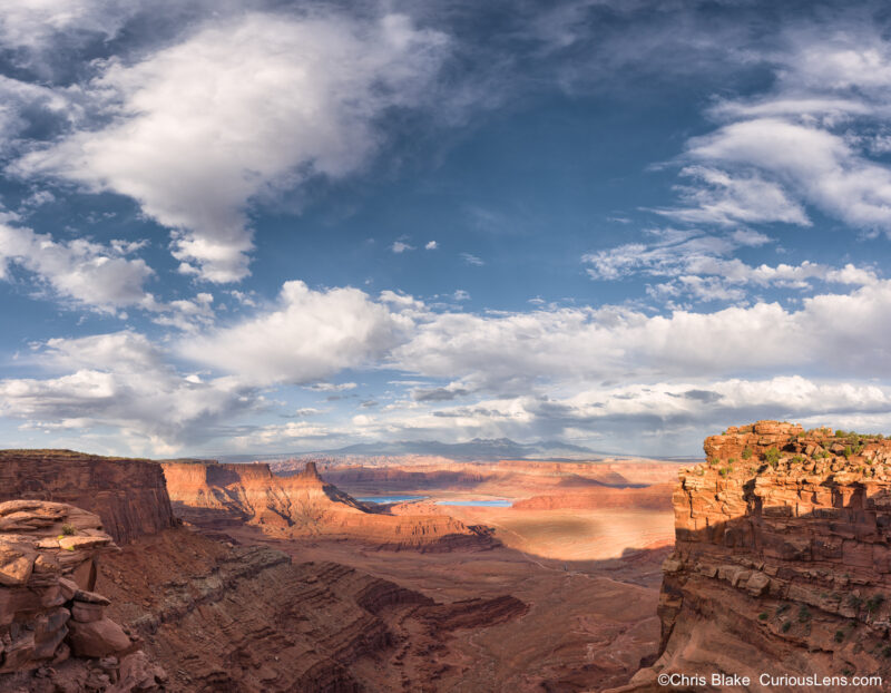 Wide panoramic view from Dead Horse Point State Park toward the La Sal Mountains, with canyon shadows, desert cliffs, storm clouds, and distant potash evaporation ponds.